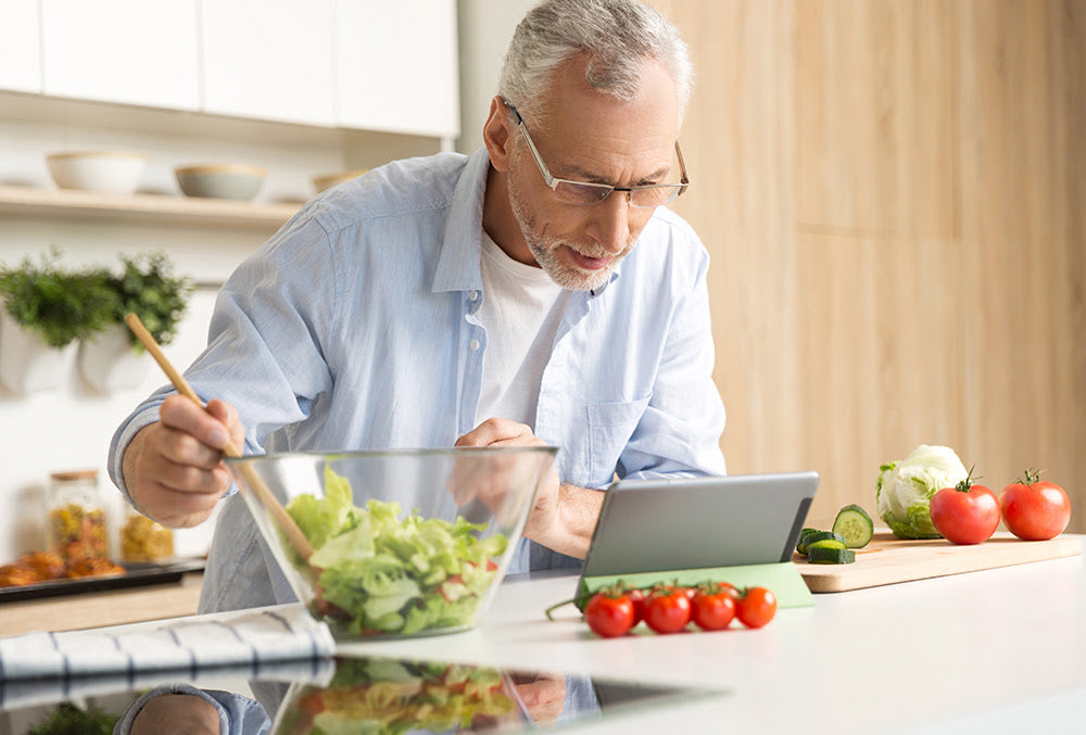 Older man in glasses preparing a fresh salad in the kitchen while looking at a tablet, focusing on healthy nutrition with vegetables on the counter.