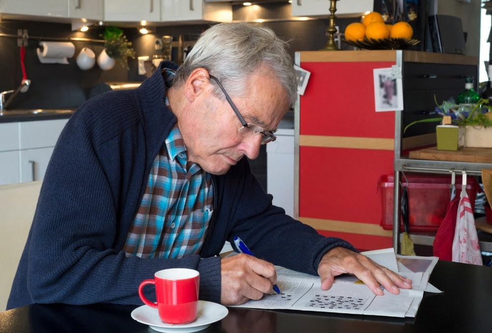 Older man wearing glasses concentrating while solving a crossword puzzle at the kitchen table, with a cup of coffee beside him, supporting mental sharpness.