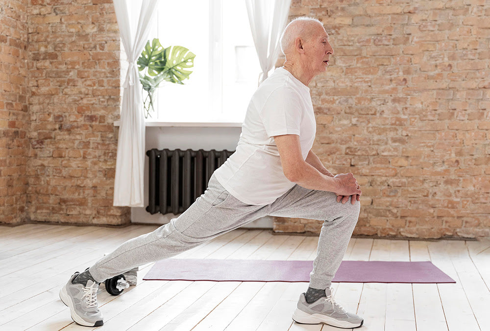 Elderly man in athletic wear performing a deep lunge stretch indoors on a yoga mat, promoting mobility and flexibility.
