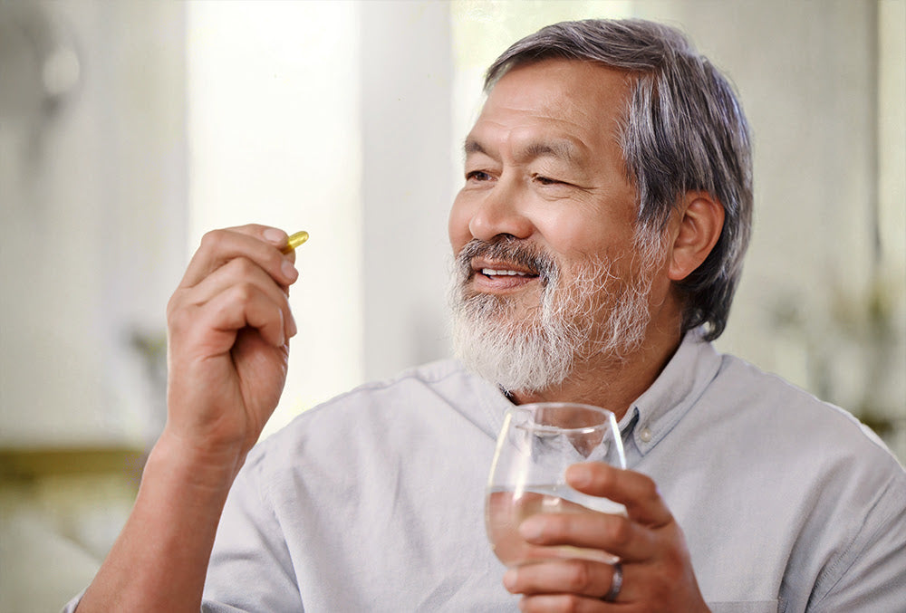 Older man with a gray beard smiling while holding a glass of water in one hand and a supplement capsule in the other, emphasizing healthy supplementation.
